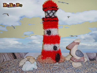 Gehäkelter rot-weißer Leuchtturm aus flauschigem Garn auf Sand neben Keramikvogel und Fischfigur