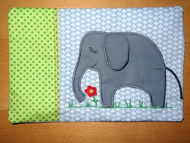 Grau applizierter Elefant mit aufgenähter Ohrklappe, gesticktem Gras und roter Blume auf hellblauem Stoff neben grüner gepunkteter Stoffbahn