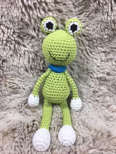 Hellgrüner Amigurumi-Frosch mit blauem Kragen und weißen Händen und Füßen auf flauschigem Hintergrund