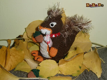 Gehäkeltes braunes Amigurumi-Eichhörnchen mit flauschigem Schwanz hält Eichel auf gelben Blättern