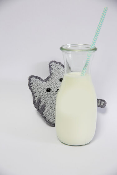 flaches graues Amigurumi-Kätzchen mit dunklem Rand und runden schwarzen Augen, das hinter einer Glasmilchflasche hervorlugt