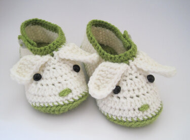 Weiß-grüne handgehäkelte Babyschuhe mit Schäfchendetails, Ohren und dunklen Knopfaugen auf hellem Hintergrund.