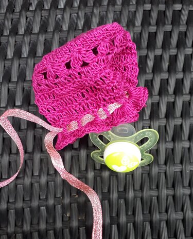 Nahaufnahme einer rosa Häkelmütze mit Banddetail neben Schnuller auf dunklem Rattan