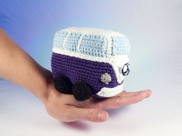 Hand hält einen gehäkelten Amigurumi-Bus in Lila, Weiß und Hellblau.