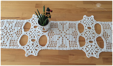 Weißer gehäkelter Tischläufer mit Blumen- und Gittermuster auf Holztisch, daneben eine kleine Pflanze.