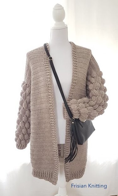 Mannequin wearing a beige open-front crochet cardigan with bubble-stitch sleeves and ribbed hem, styled with a crossbody bag.