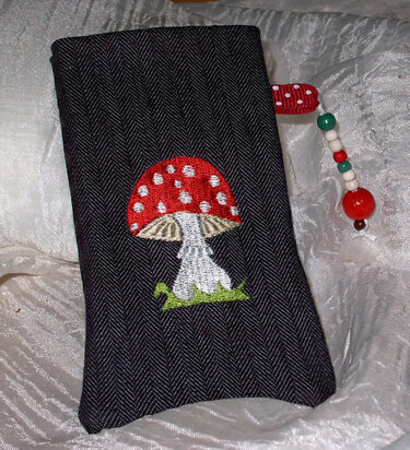 Dunkler Fischgrätstoff mit besticktem roten Fliegenpilz, grünen Grasstichen und Perlenanhänger