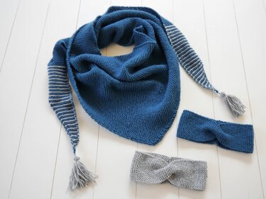 Dunkelblaues gestricktes Dreieckstuch mit gestreiften Enden und Quasten, dazu blaues und graues gedrehtes Stirnband auf hellem Boden