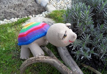 Gehäkelte Amigurumi-Schildkröte mit beigem Körper und Regenbogenpanzer auf einem Holzstück im Garten neben einem Lavendelbusch.