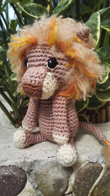 Gehäkelter Amigurumi-Löwe in Beige mit orange-gelber Mähne, sitzend auf Stein vor Pflanzen