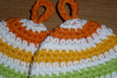 Zwei gestreifte gehäkelte Topflappen in Orange, Weiß, Gelb und Grün mit Aufhängeschlaufen auf Holz.