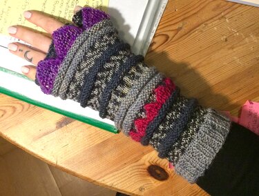 Hand mit fingerlosen, gestrickten Armstulpen in Grau mit lila Rüsche und rotem Zickzack