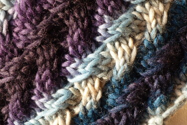 Nahaufnahme gestrickter Textur in Violett, Blau und Beige mit zopfartiger, voluminöser Optik.