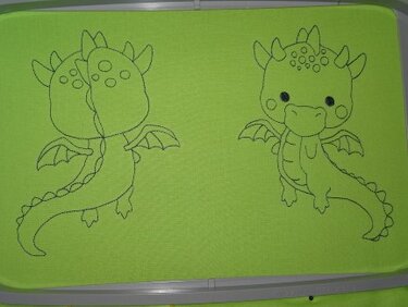 Zwei gestickte Umrisse von niedlichen Zeichentrickdrachen auf grünem Stoff im Stickrahmen