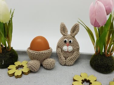 Gehäkelter Eierwärmer in Hasenform neben einem gehäkelten Eierbecher mit Pfoten und braunem Ei