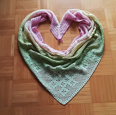 Gehäkeltes Dreieckstuch in Pastell (Rosa, Gelb, Grün) mit Lochmuster, herzförmig auf Parkett drapiert.