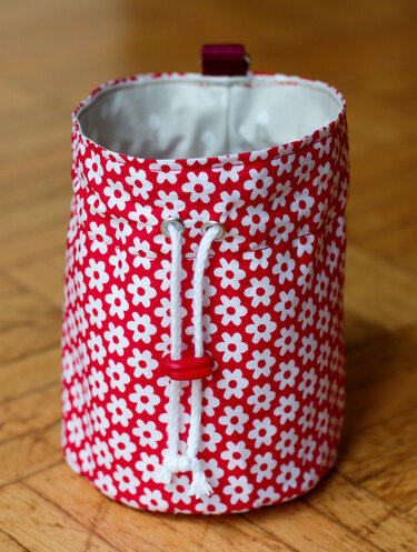 Rote Stofftasche mit weißen Blumen, Kordelzug und rotem Stopper auf Holzboden