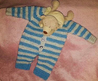 Handgehäkelter Babystrampler blau und hellgrau gestreift mit Knopfleiste auf rosa Decke, daneben ein Teddybär.