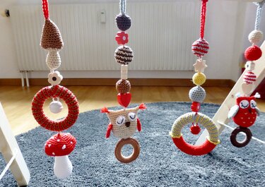 Detailansicht von vier handgehäkelten Anhängern am Baby-Spieltrapez, darunter Pilze, Eulen und Greifringe. Mit Holzperlen und Glöckchen in Naturtönen.