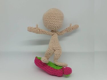 Gehäkelter Amigurumi-Puppenkörper in Beige auf pink-grünem Brett mit ausgestreckten Armen.