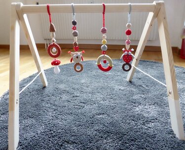 Vier gehäkelte Anhänger (zwei Eulen, ein Pilz und Greifringe) an einem minimalistischen Holz-Babyspielbogen über einem grauen Teppich.