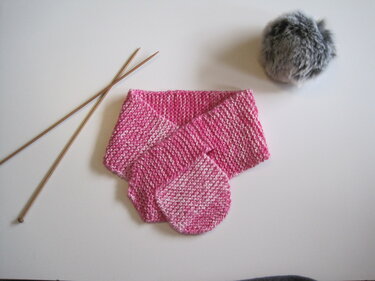 Gestrickter pinker Babyschal neben Holzstricknadeln und grauem Bommel