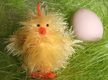 Gelbes flauschiges Strickküken mit orangem Schnabel und Füßen neben einem hellen Ei auf grünem Dekogras.