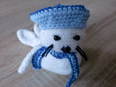 Weißer gehäkelter Seehund Amigurumi mit blauer Matrosenmütze und Schal auf Holzuntergrund