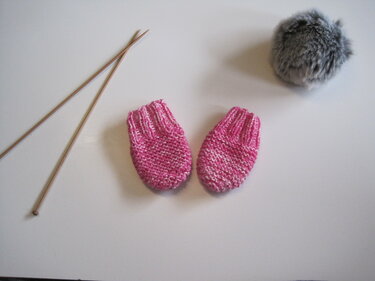 Zwei rosa‑weiß melierte, handgestrickte Babyfäustlinge neben zwei Holzstricknadeln und einem grauen Bommel auf hellem Untergrund.