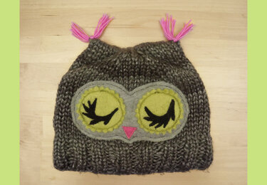 Grau-braune gestrickte Eulenmütze mit gelben Filzaugen, schwarzen Wimpern und pinken Quasten