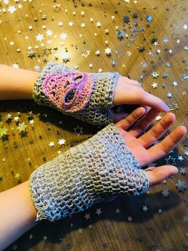 Zwei Hände tragen graue, gehäkelte, fingerlose Armstulpen; auf einem Handrücken sitzt eine rosa Totenkopf-Applikation, Hintergrund mit Sternkonfetti
