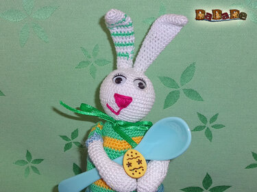Gehäkelter weißer Hase mit gestreiften Ohren, grüner Schleife, hält blauen Löffel und kleines verziertes Ei