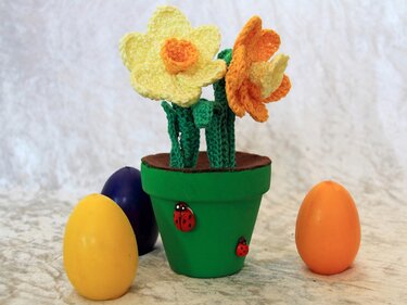 Gehäkelte gelbe und orange Narzissen in grünem Blumentopf mit drei bunten Eiern