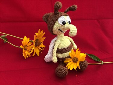 Gehäkelte Amigurumi-Biene mit braun-beigen Streifen, weißen Flügeln und Fühlern auf rotem Stoff neben gelben Blüten.