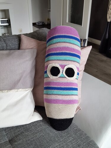 Zylindrisches gehäkeltes Kissen in Bleistiftform mit bunten Streifen und aufgenähten Augen auf einem Sofa.