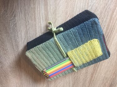 Drei gehäkelte Stiftetuis in Blau- und Grüntönen, zusammengebunden, eines mit farbigen Buntstiften, auf Holzuntergrund.