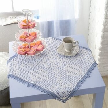 Quadratisches weißes Häkeldeckchen mit blauem Spitzenrand auf einem hellblauen Tisch, dekoriert mit einer Etagère mit Gebäck und einer Tasse.