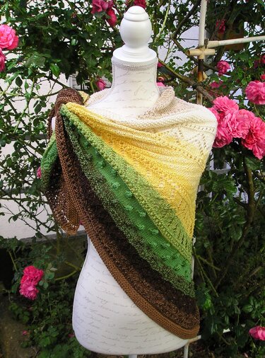 Gestricktes Dreieckstuch mit Farbverlauf in Creme, Gelb, Grün und Braun auf einer Schaufensterpuppe vor Rosen.