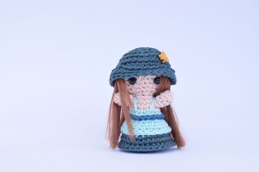 Gehäkelte Amigurumi-Puppe mit braunen Haaren, zweifarbigem blauem Kleid und dunkelblauem Hut mit gelbem Detail.