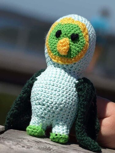 Hellblaue Amigurumi-Eule mit dunkelgrünen Flügeln und orangefarbenem Schnabel auf Holz