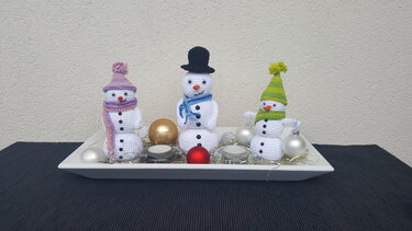Drei gehäkelte Amigurumi-Schneemänner mit Mützen und Schals auf einem dekorierten Tablett.