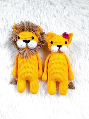 Zwei gehäkelte Löwen-Amigurumi, einer mit Mähne, einer mit Blüte, auf weißem flauschigem Hintergrund.