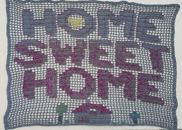 Gehäkeltes Filetbild mit Schrift 'HOME SWEET HOME', graues Netz, pink-violette Buchstaben, gelbes Herz und kleines Häuschen