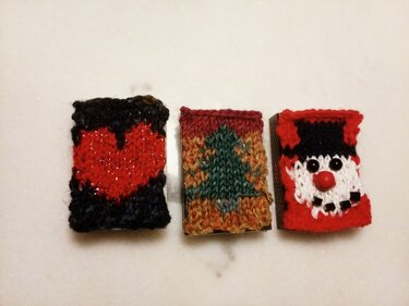 Drei gestrickte Mini‑Hüllen mit Herz-, Tannenbaum- und Weihnachtsmann‑Motiv