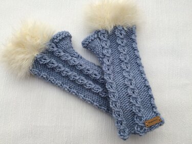 Zwei hellblaue, gestrickte fingerlose Armstulpen mit Zopfmuster und cremefarbenem, flauschigem Fellrand auf hellem Stoff.