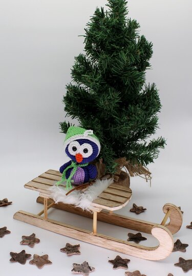 Gehäkelte Amigurumi-Eule mit grünem Mützchen auf Holzschlitten neben Mini-Tannenbaum