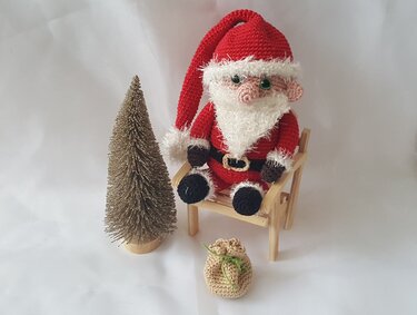 Gehäkelter Weihnachtsmann-Amigurumi in rotem Kostüm auf kleinem Holzstuhl, daneben goldene Tanne und gehäkelter Sack
