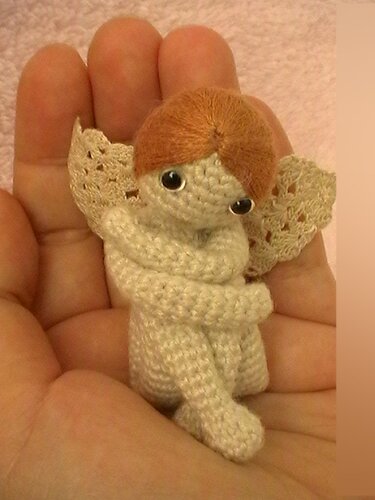 Nahaufnahme eines kleinen gehäkelten Engelchens mit verschränkten Beinen und Spitzenflügeln in einer Hand