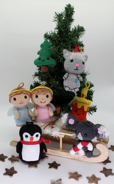 Verschiedene gehäkelte Anhänger (Engel, Pinguin, Katze, Koala, Tanne, Kerze) mit Schlaufen, arrangiert um einen Mini‑Schlitten vor einem kleinen Tannenbaum