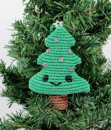 Gehäkelter grüner Weihnachtsbaum-Anhänger mit lächelndem Gesicht, rosa Perlen und kleinem Stern, an einem Tannenzweig aufgehängt